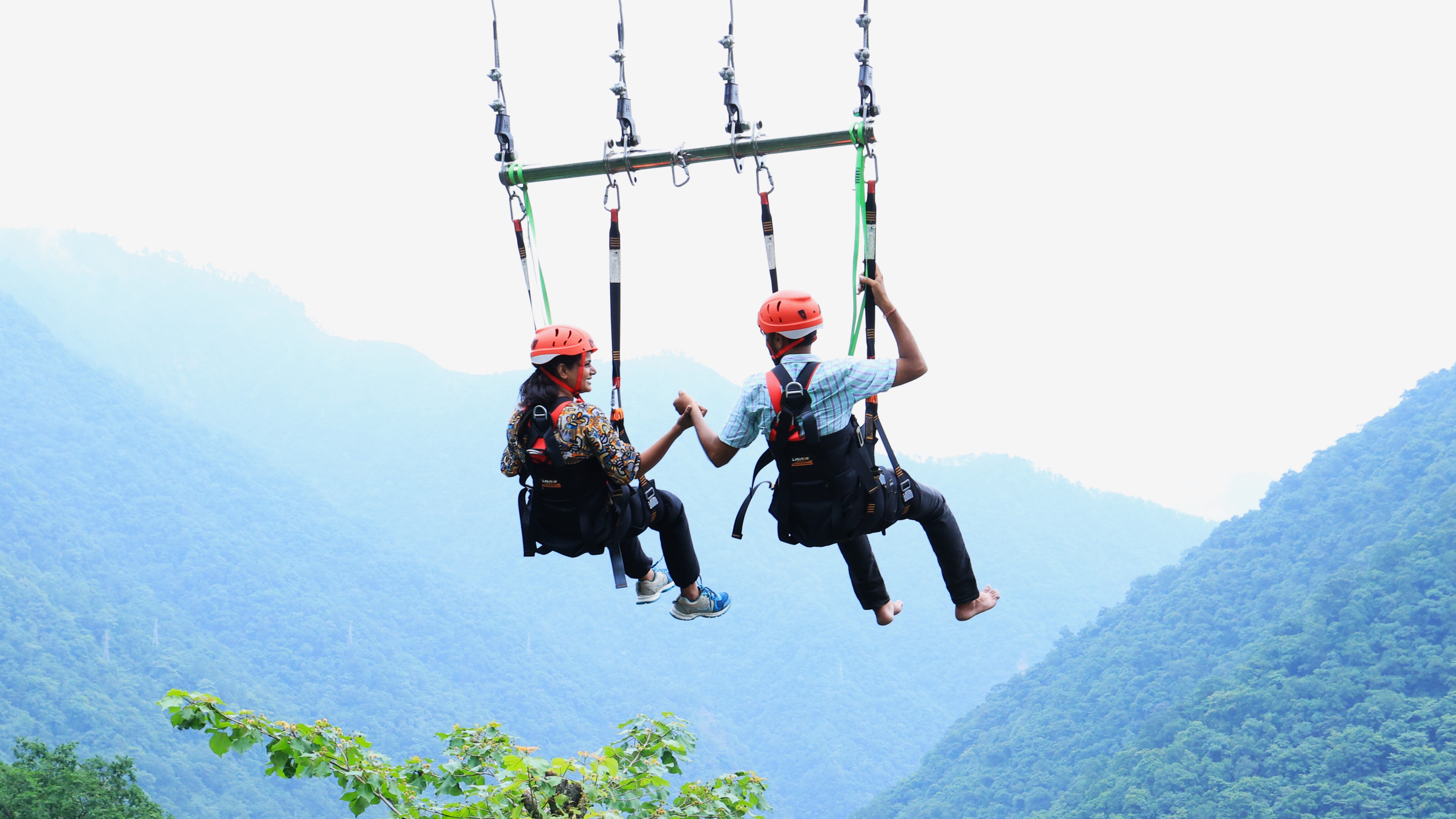 Giant swing couple activity in Rishikesh with two participants wearing safety helmets and harnesses above a green mountain valley.